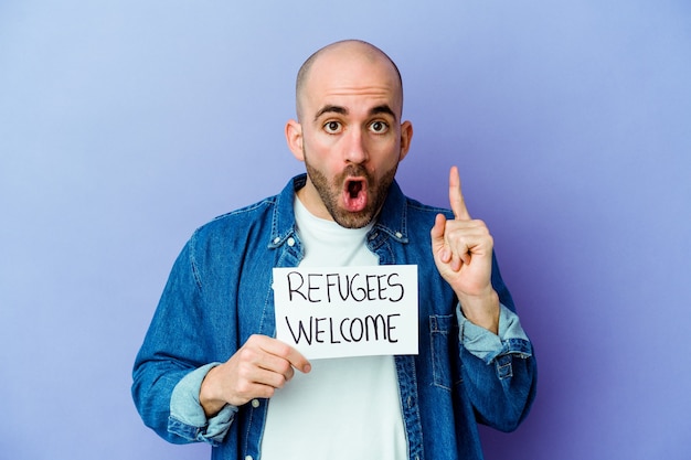 Young caucasian bald man holding a Refugees welcome placard isolated on blue wall having an idea, inspiration concept.