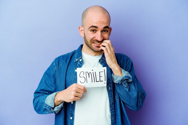 Young caucasian bald man holding a Smile placard isolated on purple wall biting fingernails, nervous and very anxious.