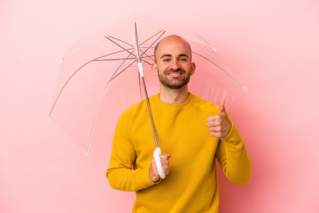 Молодой кавказский лысый мужчина держит зонтик на розовом фоне, улыбаясь и поднимая палец вверх