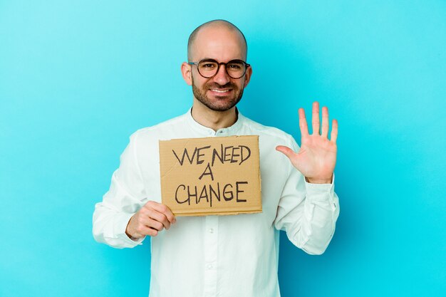 Молодой кавказский лысый мужчина держит табличку с надписью «Нам нужно изменение» на фиолетовом фоне, улыбаясь, весело показывая номер пять с пальцами.