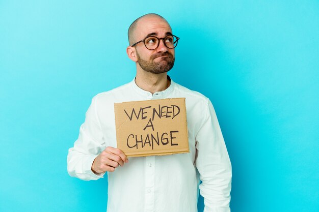 Young caucasian bald man holding a We need a change placard isolated on purple wall dreaming of achieving goals and purposes