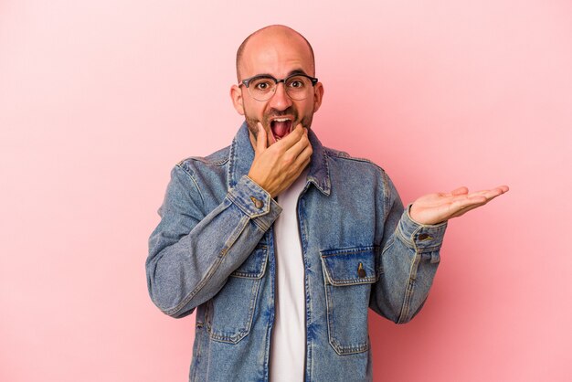 Young caucasian bald man isolated on pink background  holds copy space on a palm, keep hand over cheek. Amazed and delighted.