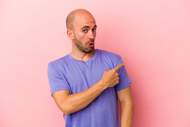 Young caucasian bald man isolated on pink background  smiling and pointing aside, showing something at blank space.