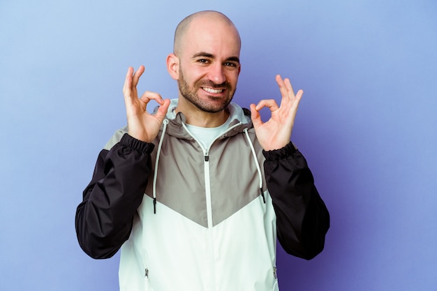 Young caucasian bald man isolated on purple background cheerful and confident showing ok gesture.