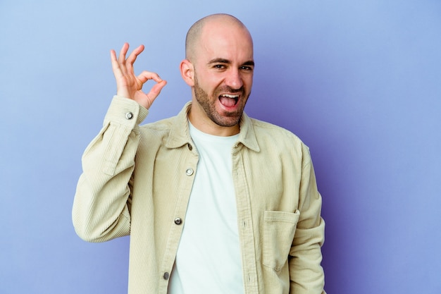 Young caucasian bald man isolated on purple background winks an eye and holds an okay gesture with hand.