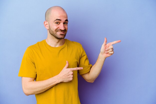 Young caucasian bald man isolated on purple wall excited pointing with forefingers away.