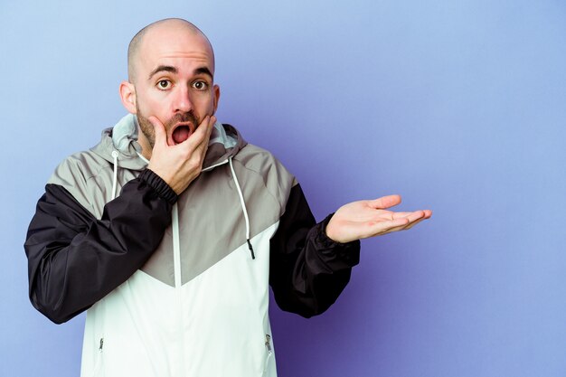 Young caucasian bald man isolated on purple wall holds copy space on a palm, keep hand over cheek. Amazed and delighted.