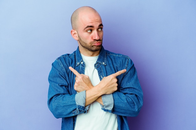 Young caucasian bald man isolated on purple wall points sideways, is trying to choose between two options.