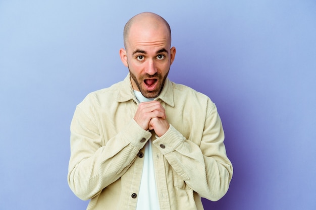 Young caucasian bald man isolated on purple wall praying for luck, amazed and opening mouth looking to front