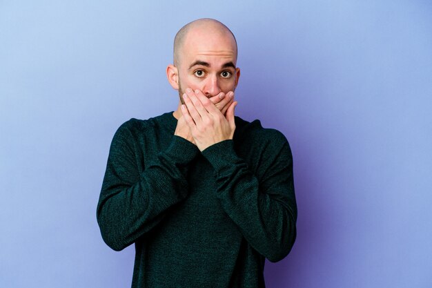 Young caucasian bald man isolated on purple wall shocked covering mouth with hands.