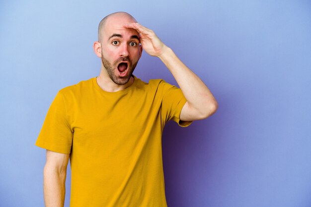 Young caucasian bald man isolated on purple wall shouts loud, keeps eyes opened and hands tense.