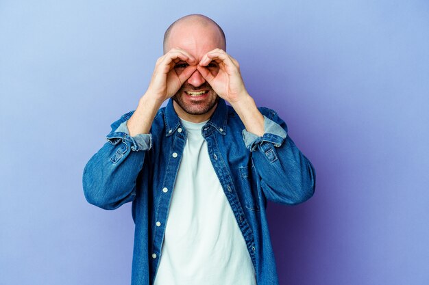 Young caucasian bald man isolated on purple wall showing okay sign over eyes