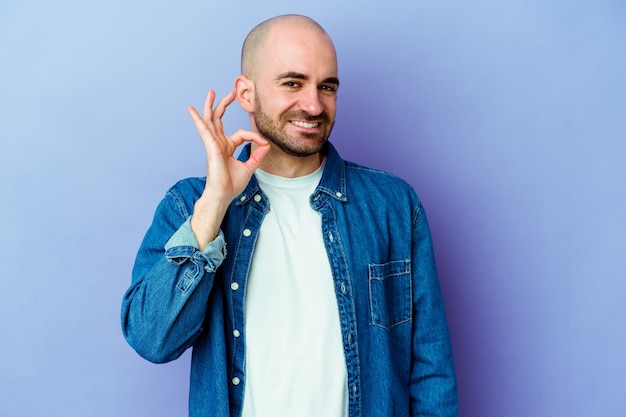 Young caucasian bald man isolated on purple wall winks an eye and holds an okay gesture with hand