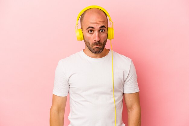 Young caucasian bald man listening to music isolated on pink background  shrugs shoulders and open eyes confused.