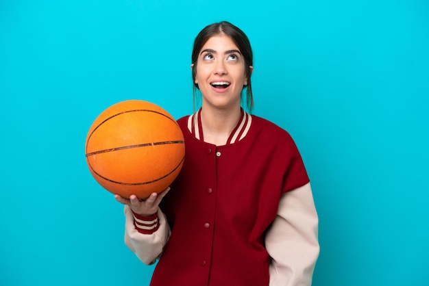 Young caucasian basketball player woman isolated on blue background looking up and with surprised expression