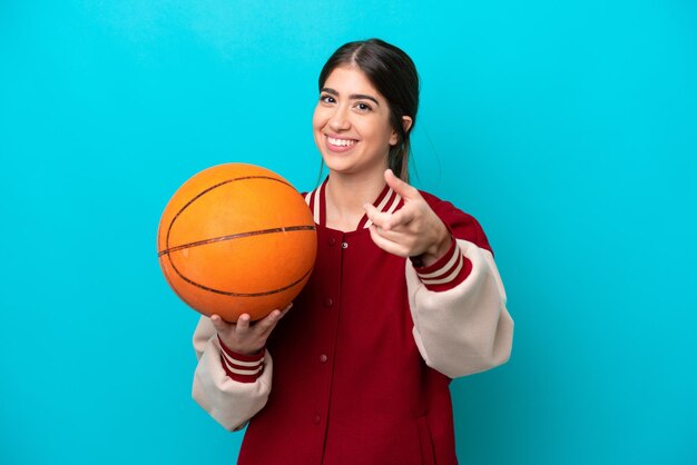 Молодая кавказская баскетболистка женщина изолирована на синем фоне, указывая вперед со счастливым выражением лица