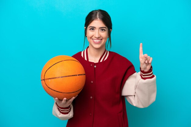 最高のサインで指を示して持ち上げて青い背景で隔離の若い白人バスケットボール選手の女性