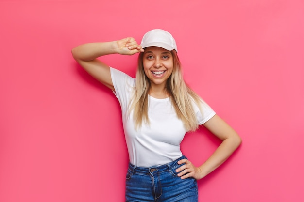 A young caucasian beautiful happy blonde woman in a white tshirt cap and blue jeans stands and smiles holding on to the visor of her cap isolated on a bright color pink wall