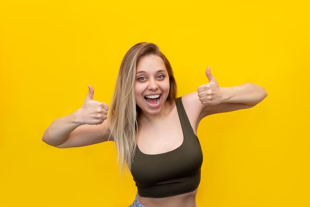 A young caucasian beautiful happy excited delighted smiling blonde woman in a short top showing thumb up like gesture with two hands isolated on a bright color yellow wall