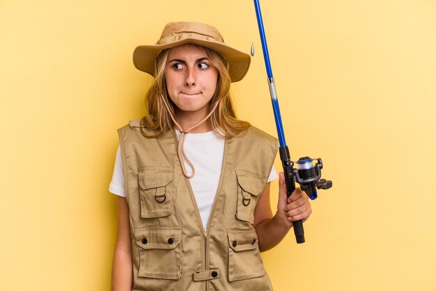 Young caucasian blonde fisherwoman holding a rod isolated on yellow background  confused, feels doubtful and unsure.