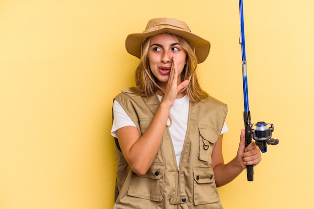 Young caucasian blonde fisherwoman holding a rod isolated on yellow background  is saying a secret hot braking news and looking aside