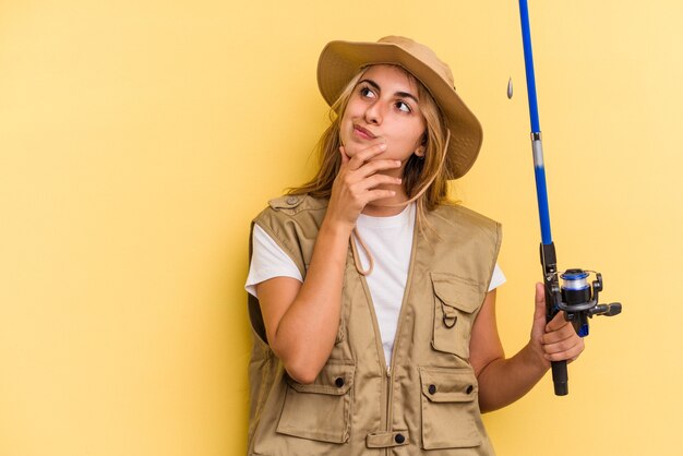 Young caucasian blonde fisherwoman holding a rod isolated on yellow background  looking sideways with doubtful and skeptical expression.