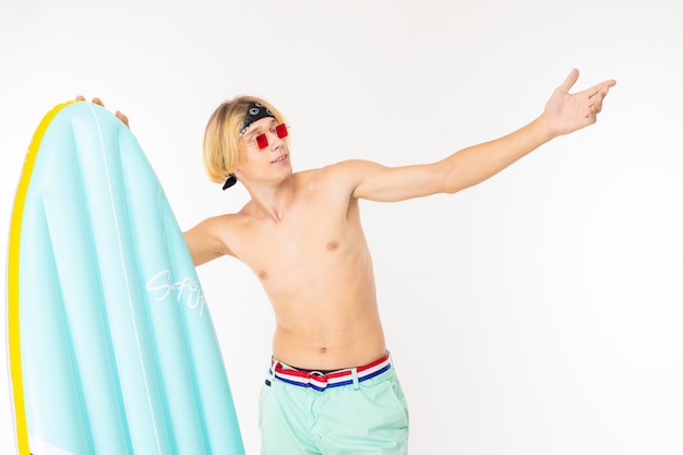 Young caucasian blonde man stands in swimsuit with big rubber beach mattress isolated on white wall