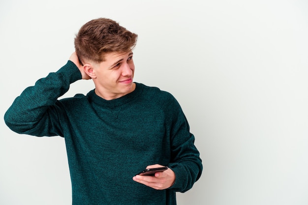 Young caucasian blonde man using a telephone isolated on white wall touching back of head, thinking and making a choice