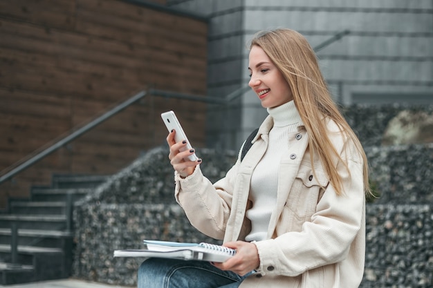 若い白人の金髪の学生の女の子が携帯電話でビデオ通話をし、大学の建物の近くのベンチに座りながら微笑む