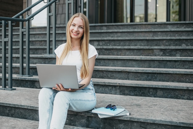 Young caucasian blonde student girl sitting with laptop on stairs near university smiling and learn English. Copy space