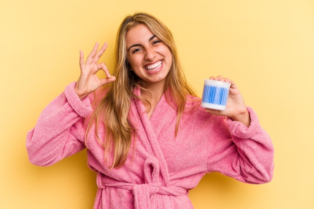 Young caucasian blonde woman holding cotton buds isolated on yellow background  cheerful and confident showing ok gesture.