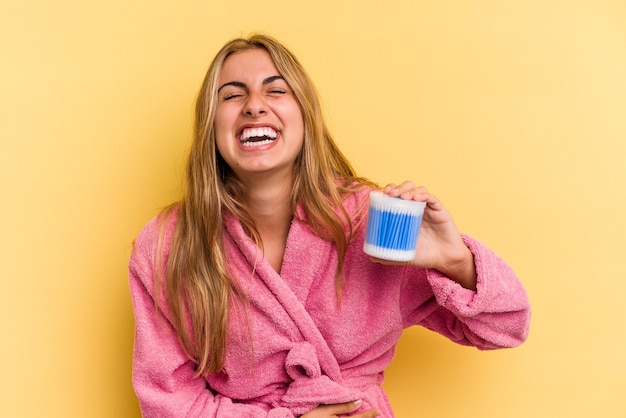 Young caucasian blonde woman holding cotton buds isolated on yellow background  laughing and having fun.