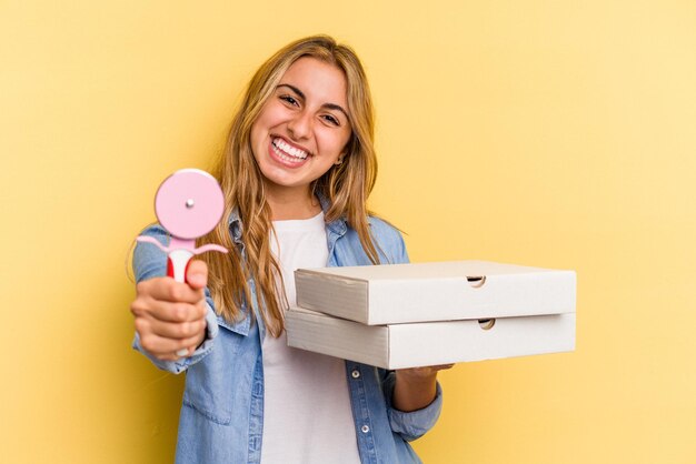 Young caucasian blonde woman holding pizzas and cutter isolated on yellow background