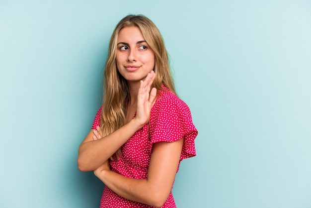 Young caucasian blonde woman isolated on blue background  confused, feels doubtful and unsure.