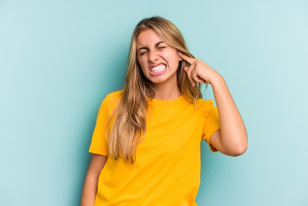 Young caucasian blonde woman isolated on blue background  covering ears with fingers, stressed and desperate by a loudly ambient.