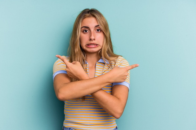 Young caucasian blonde woman isolated on blue background  points sideways, is trying to choose between two options.