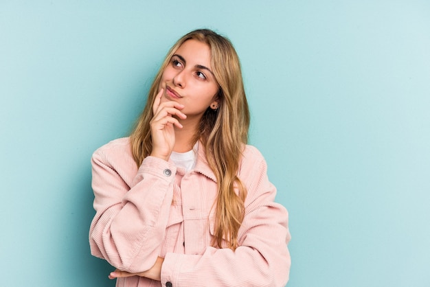 Young caucasian blonde woman isolated on blue background  thinking and looking up, being reflective, contemplating, having a fantasy.