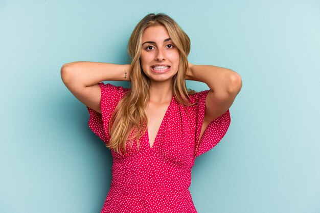 Young caucasian blonde woman isolated on blue background  touching back of head, thinking and making a choice.