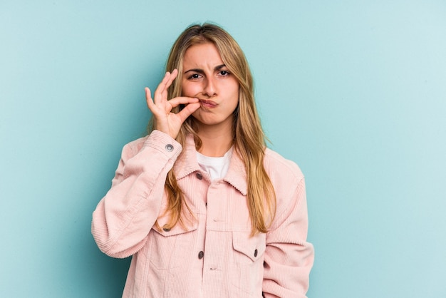 Young caucasian blonde woman isolated on blue background  with fingers on lips keeping a secret.