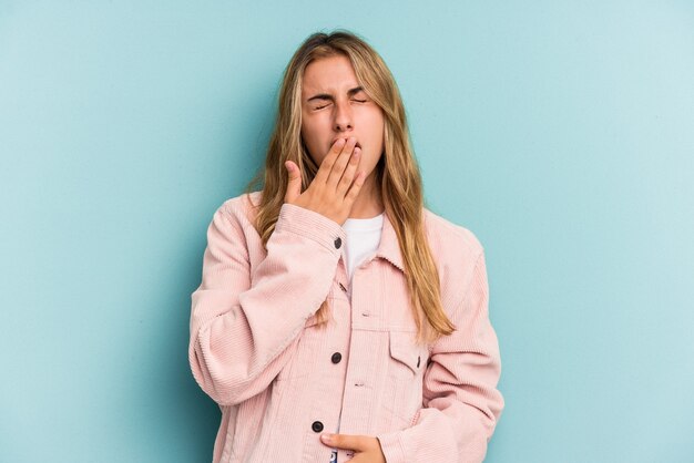 Young caucasian blonde woman isolated on blue background  yawning showing a tired gesture covering mouth with hand.