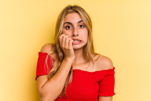 Young caucasian blonde woman isolated on yellow background  biting fingernails, nervous and very anxious.