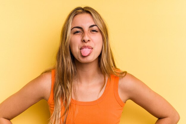 Young caucasian blonde woman isolated on yellow background  funny and friendly sticking out tongue.