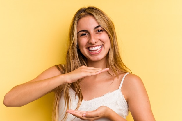 Young caucasian blonde woman isolated on yellow background holding something with both hands, product presentation