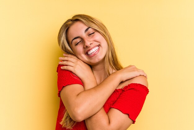 Young caucasian blonde woman isolated on yellow background  hugs, smiling carefree and happy.