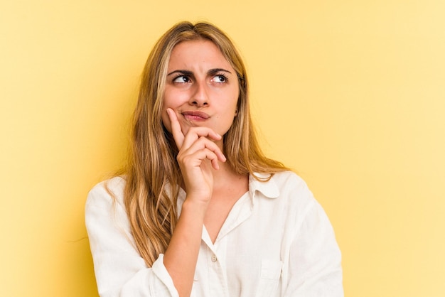 Young caucasian blonde woman isolated on yellow background  looking sideways with doubtful and skeptical expression.