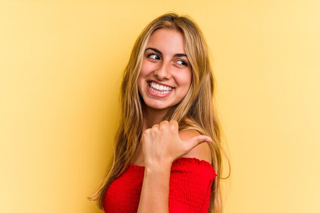 Young caucasian blonde woman isolated on yellow background  points with thumb finger away, laughing and carefree.