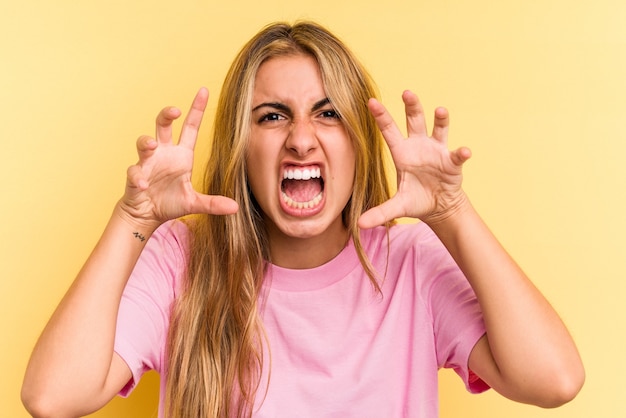 Young caucasian blonde woman isolated on yellow background  showing claws imitating a cat, aggressive gesture.