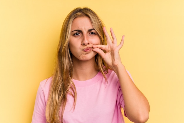 Giovane donna bionda caucasica isolata su sfondo giallo con le dita sulle labbra mantenendo un segreto.