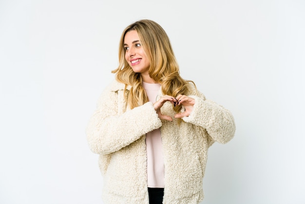 Young caucasian blonde woman smiling and showing a heart shape with hands.