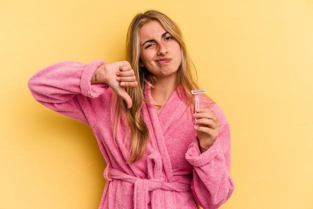 Young caucasian blonde woman wearing bathrobe holding blades isolated on yellow background  showing a dislike gesture, thumbs down. Disagreement concept.